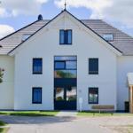 Weißes Haus mit dunklen Fenstern und Dach, Vorgarten mit Bank und Holzgarage.