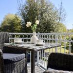 Terrasse mit Tisch, zwei Stühlen und Blumen. Blick auf grüne Bäume und ein Haus.