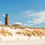 Roter Leuchtturm auf sandigem Strand mit Dünen und blauem Himmel.