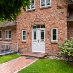 Rotes Backsteinhaus mit Strohdach, weißer Eingangstür und gepflasterter Weg.