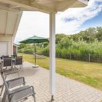 Terrasse mit Tisch, Stühlen und Sonnenschirm. Grünflächen und Bäume im Hintergrund.