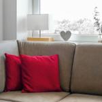Ein grauer Sofa mit zwei roten Kissen. Auf dem Fenster steht eine Lampe und ein Herz.