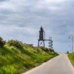 Eine Straße führt zu einem Leuchtturm auf einer grünen Anhöhe unter bewölktem Himmel.