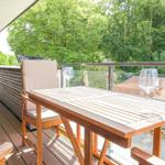 Terrasse mit Tisch, Stühlen und Weingläsern. Blick auf grüne Bäume und Dachziegel.