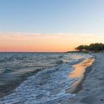 Strand mit Wellen, Sand und Wald am Horizont bei Sonnenuntergang.
