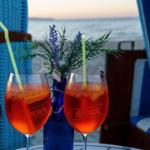 Zwei Aperol Spritz Getränke auf einem Tisch mit Meerblick und blauen Liegestühlen.