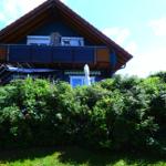 Haus mit Balkon, Blumenhecke und Sonnenschirm. Katzensilhouette auf Dach.