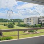 Balkon mit Sicht auf Grünfläche, Gebäude und Riesenrad unter blauem Himmel.