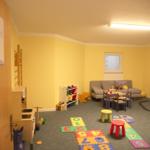 Spielzimmer mit Kindersitzgruppe, Spielzeug und Zahlenmatte auf dem Boden.