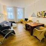 Wohn- und Essbereich mit Sofa, Esstisch und Stühlen. Holzboden und Fenster mit Vorhängen.