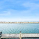 Holzsteg mit Blick auf ruhiges Wasser und Steinufer unter blauem Himmel.