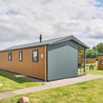 Moderne Holz-Hütte mit Terrasse und Garten. Dach mit Schornstein. Hintergrund mit Bäumen.
