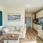 Wohnbereich mit Sofa, Couchtisch und Küchenbereich. Blaue Vorhänge und Holzboden.