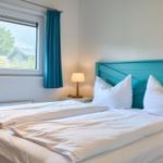 Schlafzimmer mit Doppelbett, blauem Kopfteil und Fenster mit Blick auf Grün.