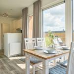 Essbereich mit Tisch und Stühlen vor großem Fenster. Küche und Wohnbereich sichtbar.