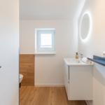 Badezimmer mit Waschbecken, Spiegel und Toilette. Holzboden und Fenster.