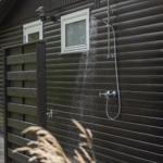 Außen-Dusche an dunklem Holzhaus mit Fenster und Pflanzen im Vordergrund.