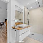 Badezimmer mit Dusche, Waschtisch und Spiegel. Durchgang in Wohnbereich mit Esstisch.