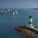 Leuchtturm mit grün-weißem Gestell steht auf Steinküste. Segelboote segeln im Hafen.