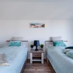 Zwei Betten mit hellblauen Bettwäsche und Kissen. Zwischen ihnen steht ein Nachttisch mit Lampe und Pflanzen.