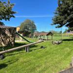 Großer Spielplatz mit Holzspielgeräten, Rutsche und Schaukel auf grünem Rasen.