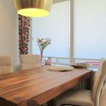 Esstisch mit Stühlen vor Fenster mit Jalousien und Blumenvase.