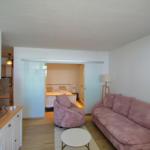 Wohnbereich mit Sofa, Sessel und TV. Schlafzimmer dahinter durch Glaswand abgetrennt.