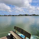 Holzbank am Wasserufer mit Blick auf Häuser am gegenüberliegenden Ufer.