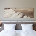 Doppelbett mit weißen Kissen und Kopfteil. Wandbild zeigt Strand mit Holzweg.