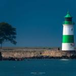 Leuchtturm mit grün-weißem Muster steht am Wasser. Hinter ihm Bäume und steinerner Wall.