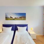 Zwei Betten mit weißen Laken, blauer Kopfteilwand und Fenster mit bunten Vorhängen.