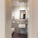 Badezimmer mit Waschbecken, Spiegel und Toilette. Graue Fliesen und weiße Wand.