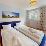 Zwei Einzelbetten mit weißen Laken, Fenster mit bunten Vorhängen und Wandbild mit Kühen.