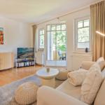 Wohnzimmer mit Sofa, Couchtisch und TV. Balkon mit Sitzgelegenheit. Fenster mit Vorhängen.