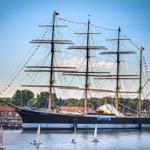Großes Segelschiff mit drei Masten liegt im Hafen. Kleine Segelboote umgeben es.
