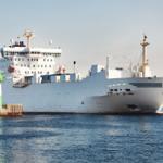 Ein großes weißes Frachtschiff fährt an einem Hafen mit Leuchtturm vorbei.