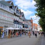 Straße mit Geschäften, Menschen und Fahrrädern unter Bäumen und bunten Girlanden.