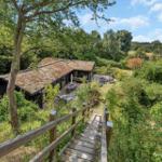 Holzsteg führt zu einem Haus mit Grasdach und Terrasse im Grünen.