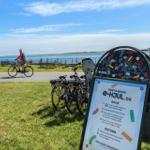 E-Bikes zum Mieten an der Wasserseite mit Fahrradweg und Bootshafen im Hintergrund.