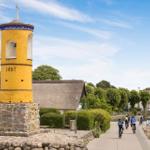 Gelber Turm mit Jahreszahl 1857, Radfahrer entlang eines Weges mit strohgedeckten Häusern.