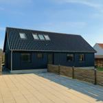 Modernes Haus mit dunklem Holz und Dachfenstern. Terrasse und Holzzaun im Vordergrund.