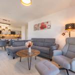 Wohnbereich mit Sofa, Sessel und Küchenbereich im Hintergrund.