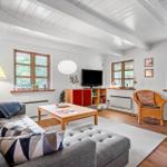 Wohnzimmer mit Sofa, Couchtisch und Fernseher. Holzdielen und große Fenster.