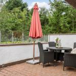 Terrasse mit Tisch, vier Stühlen und geschlossenem Sonnenschirm. Hintergrund: weiße Mauer und Bäume.