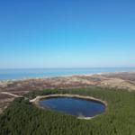Ein kleiner See liegt inmitten eines Waldes, umgeben von Hügeln und dem Meer.