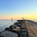 Steinpromenade am Meer bei Sonnenuntergang mit Leuchtturm und Boot.