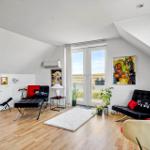 Wohnraum mit Holzboden, schwarzen Sitzmöbeln und roten Akzenten, Blick auf Wasser.