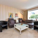 Wohnzimmer mit braunem Sofa, zwei Sesseln und Glas-Tisch. Wand mit Tapete und Kunstwerken.