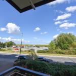 Blick von Balkon auf Straße, Parkplatz und grüne Bäume unter blauem Himmel mit weißen Wolken.
