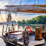 Vorderdeck eines Segelschiffs mit Ruder und Rettungsring am Wasser.